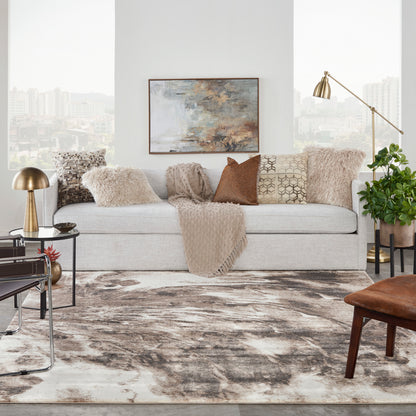 Modern area rug in light gray with an abstract pattern (9' x 12') on a living room floor, paired with a light sofa, beige throws, earthy pillows, a gold floor lamp, and a plant.