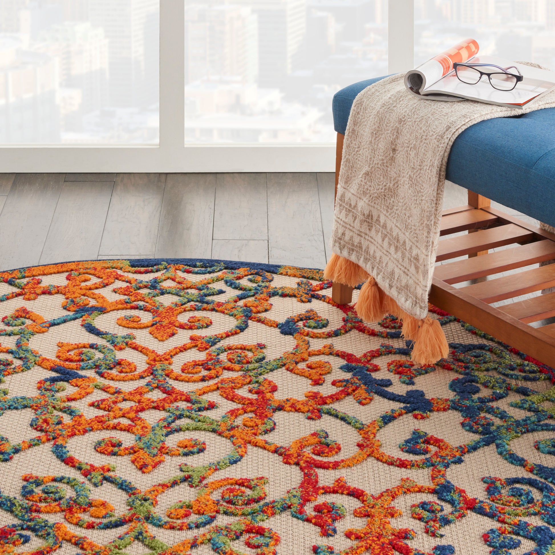 Colorful ornate area rug with orange, teal and blue swirls beside a blue bench; beige fringed throw, magazines and glasses.