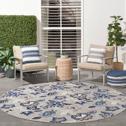 Round outdoor rug with blue floral pattern, beige patio chairs with striped cushions, wicker side table, potted plants.