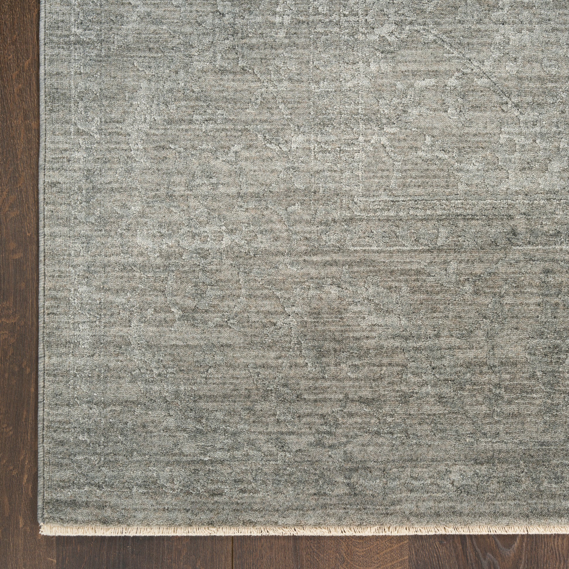 Rectangular grey textured rug with subtle pattern and light fringe along the bottom edge on dark wood floor.