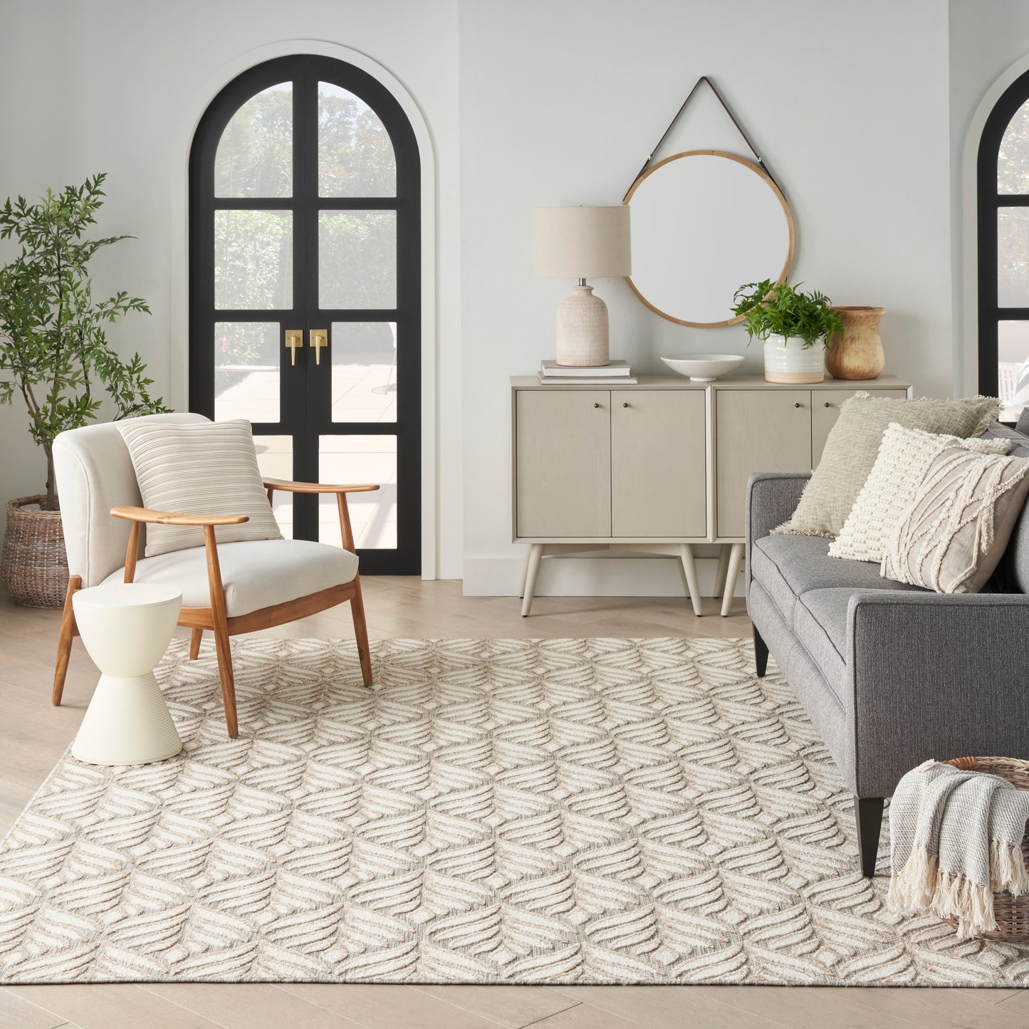 Neutral living room with arched black doors, beige woven rug, wood-armchair, gray sofa, round mirror, and sideboard decor.