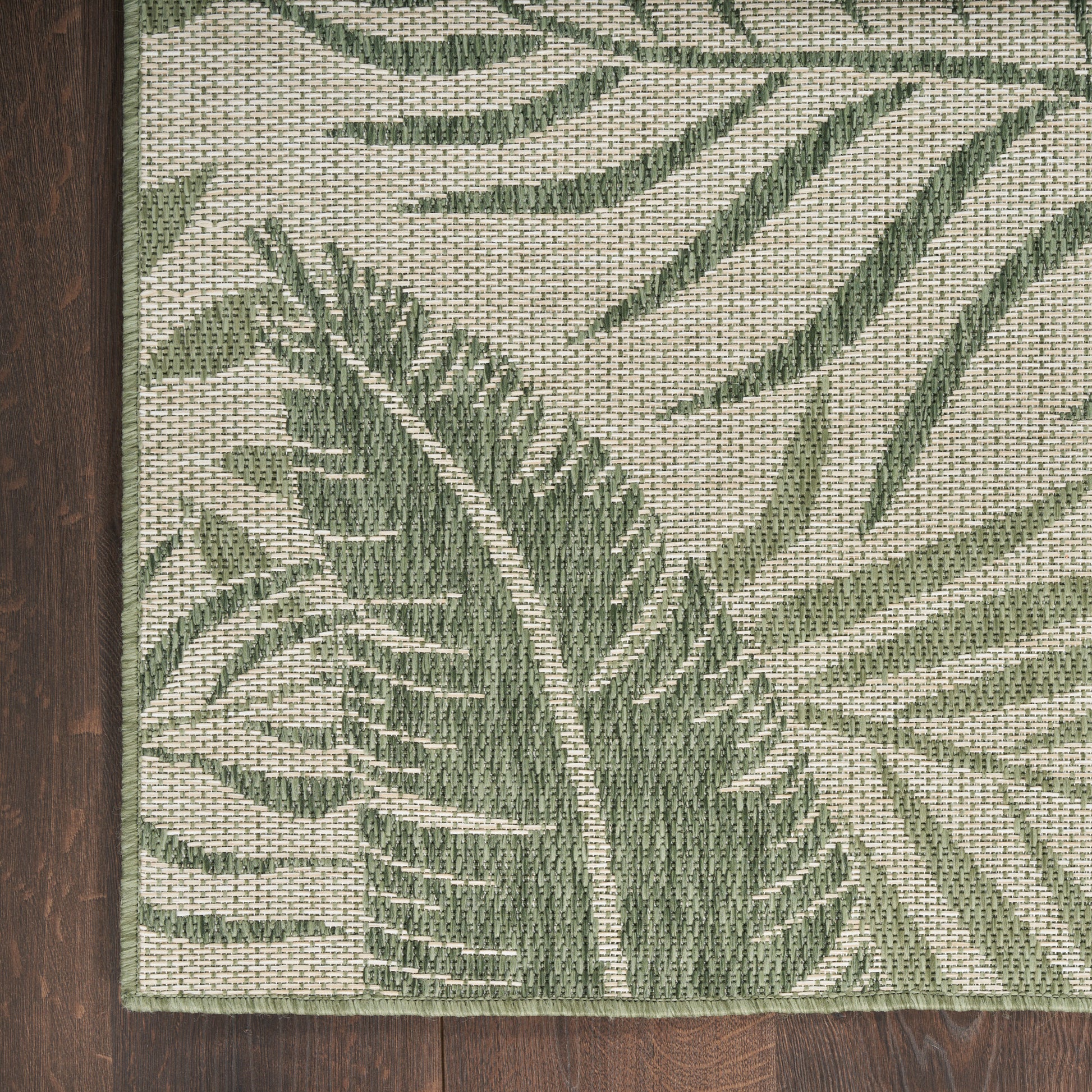 Rectangular woven rug in beige with green tropical palm leaf pattern, shown on dark wood floor.