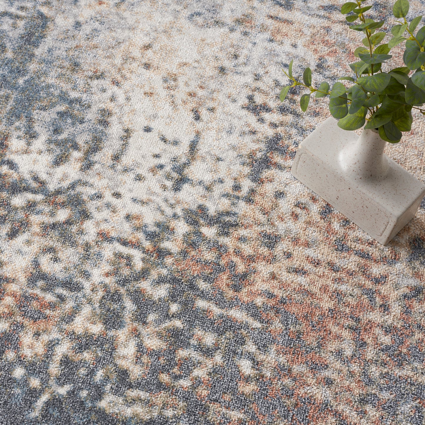Beige ceramic vase with green sprigs rests on a round abstract patterned area rug in cream, blue and rust.