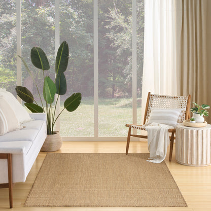 Bright living room with a beige sofa, woven rattan chair, natural jute rug, a large leafy plant, a round wood side table, and cream curtains.