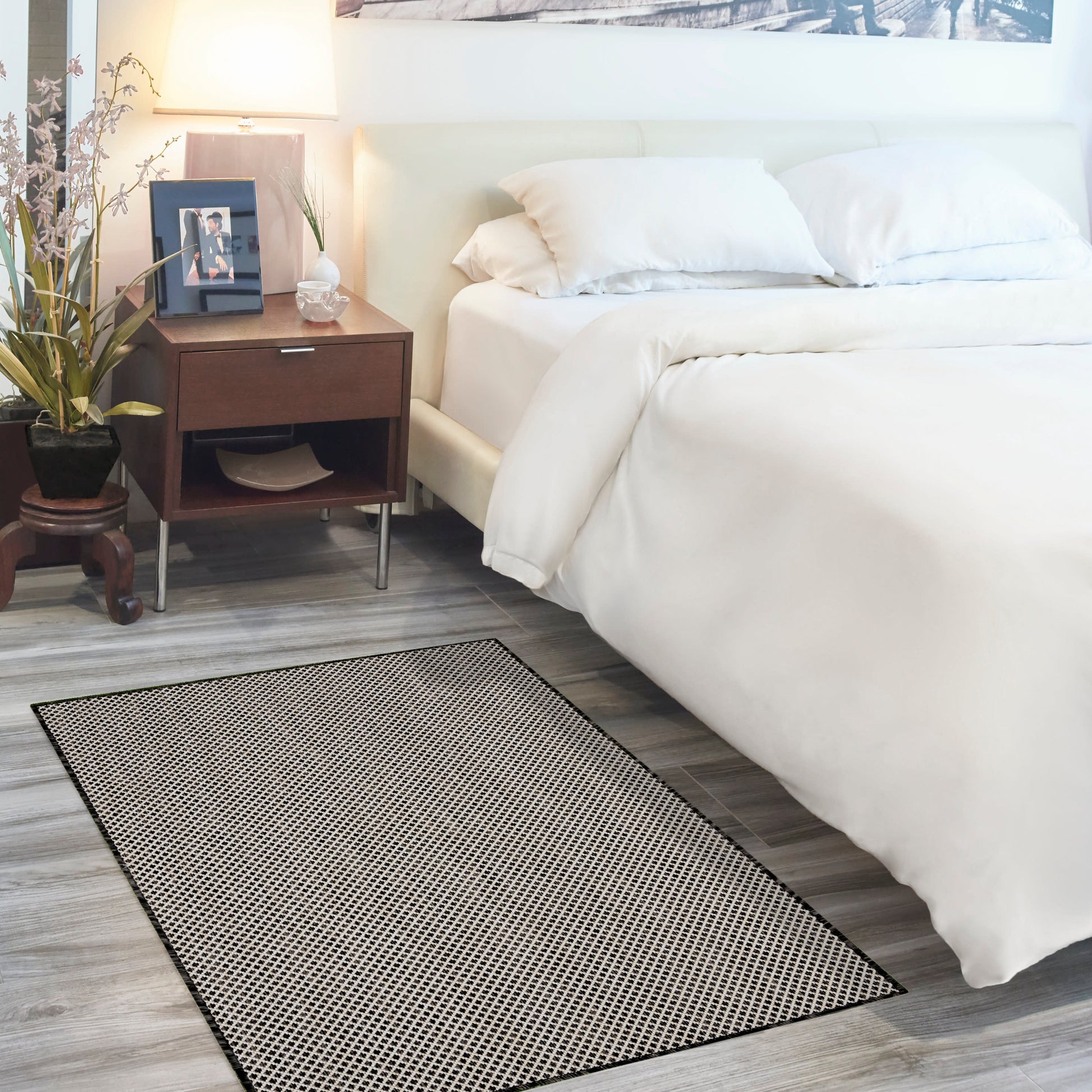 Rectangular black-dotted area rug on grey wood floor beside a white bed with a dark wood nightstand and plant.