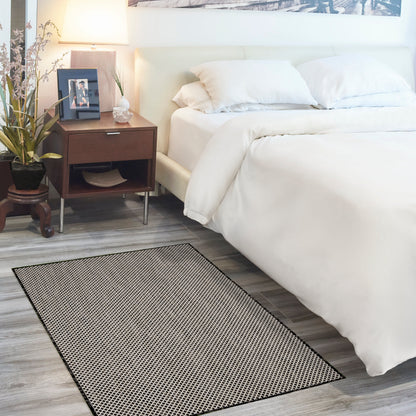 Rectangular black-dotted area rug on grey wood floor beside a white bed with a dark wood nightstand and plant.