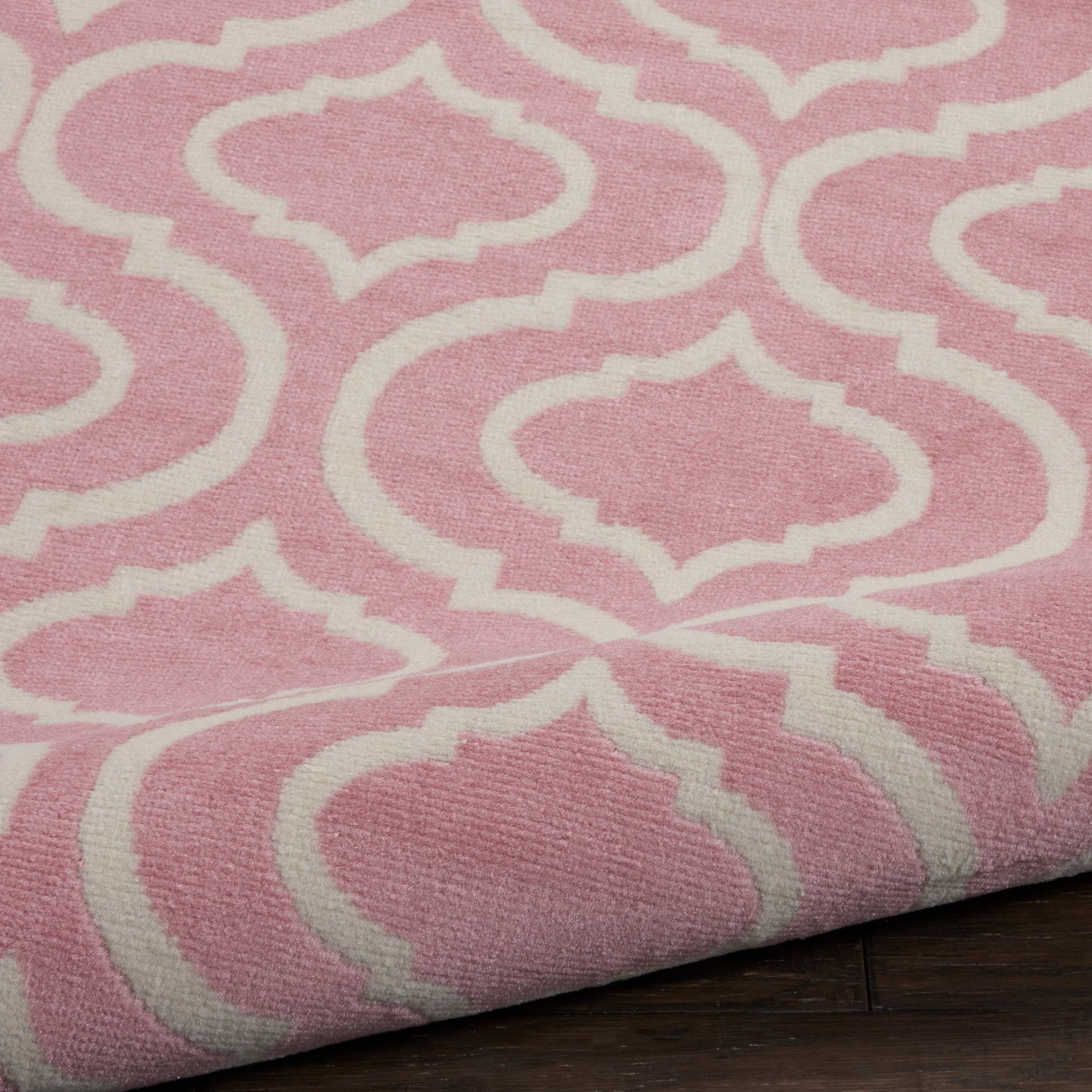 Pink woven round rug with white trellis pattern; folded edge visible on dark wooden floor.