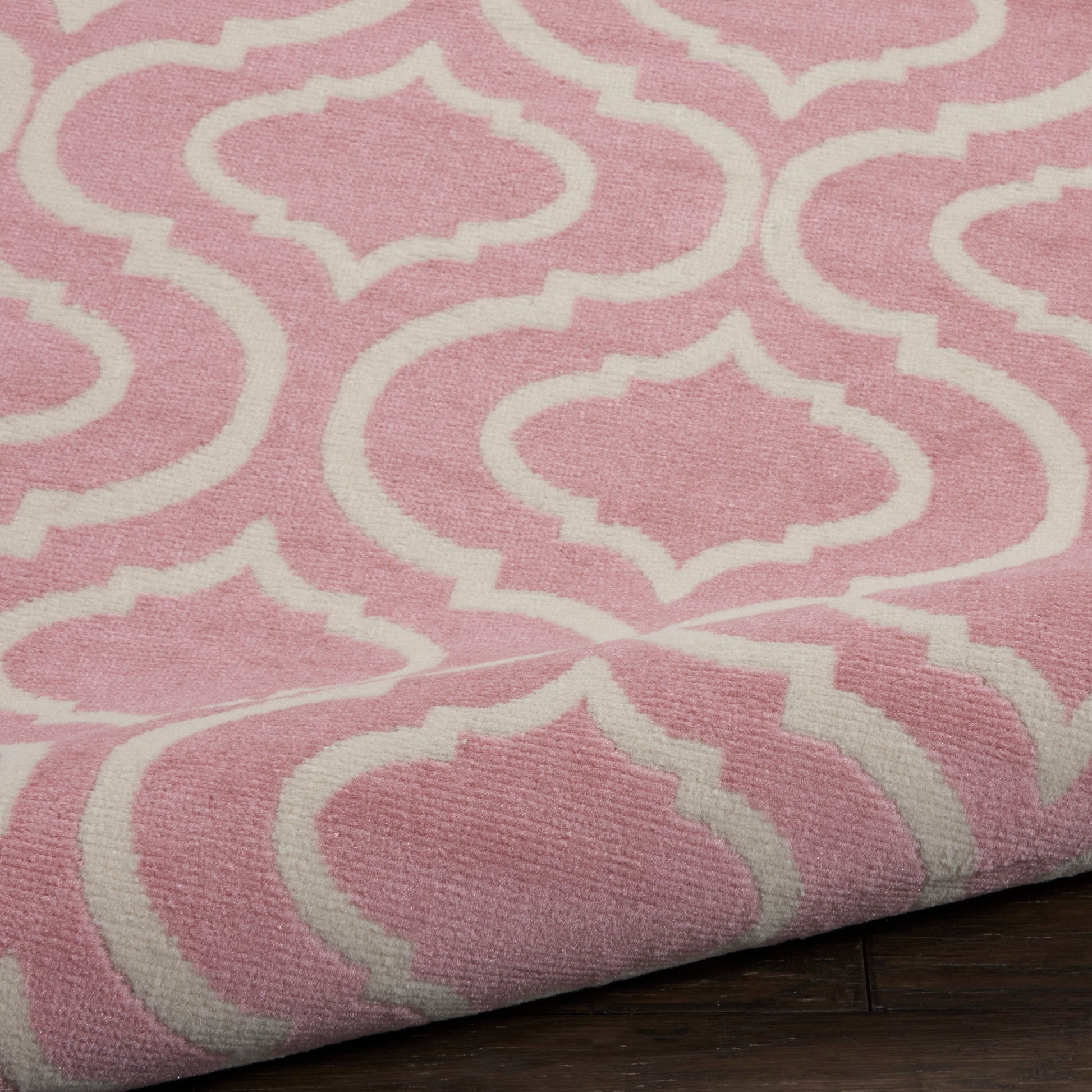 Pink woven round rug with white trellis pattern; folded edge visible on dark wooden floor.