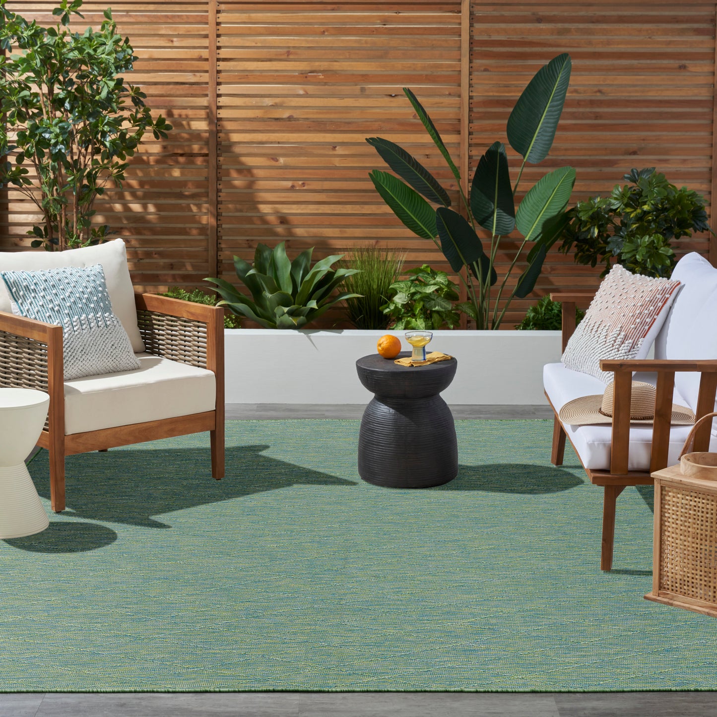 Outdoor patio scene with a teal rug, wood-framed armchairs with white cushions, woven accents, a black sculptural side table, and tropical plants.