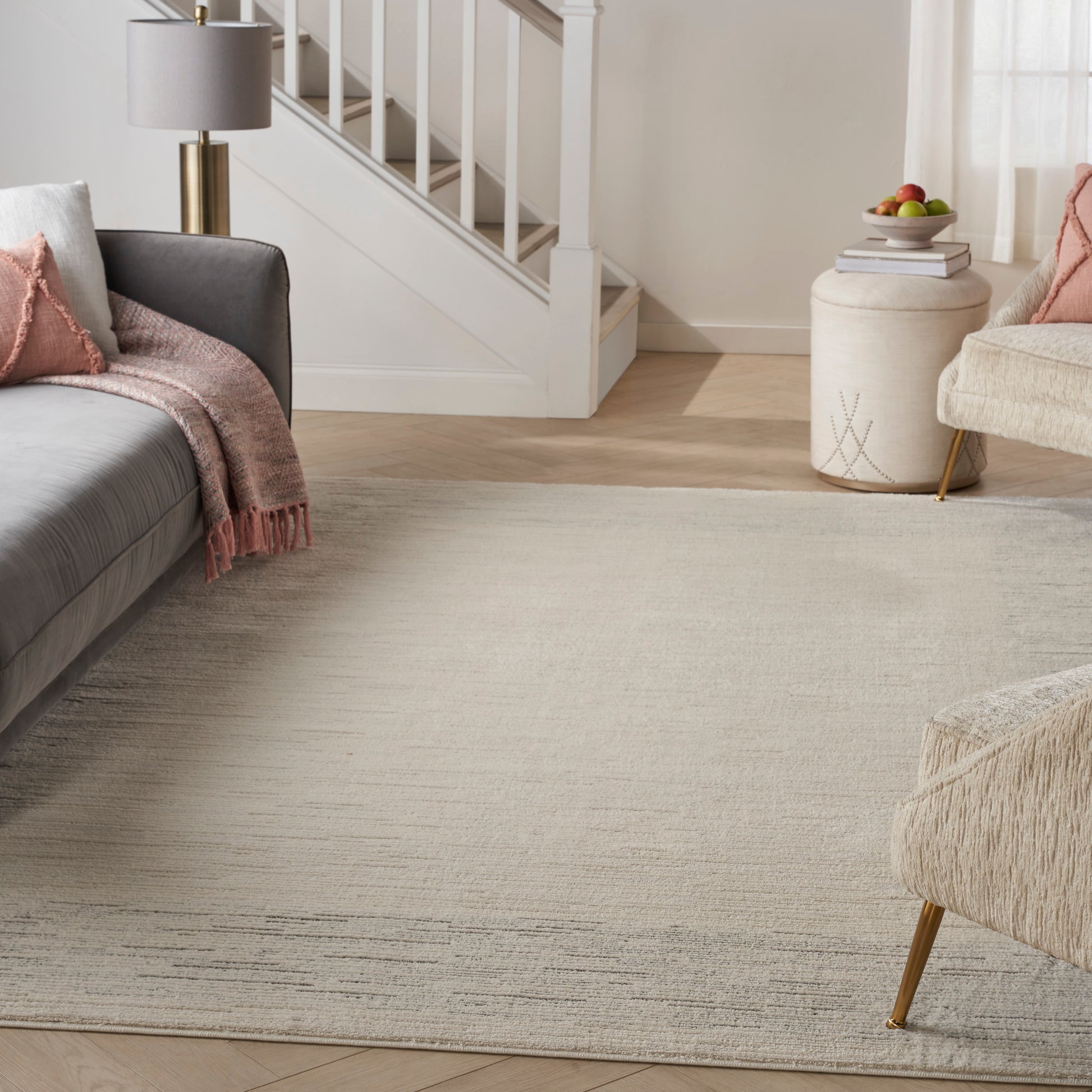 Neutral living room with gray sofa, pink throw, beige rug, light wood floor, brass lamp, round pouf by stairs.