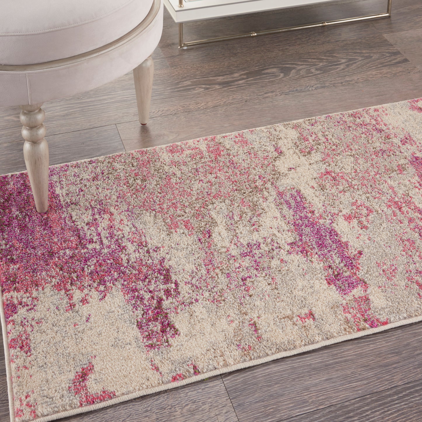 Rectangular area rug with abstract pink and purple splashes on cream, modern design beside beige chair and glass table.