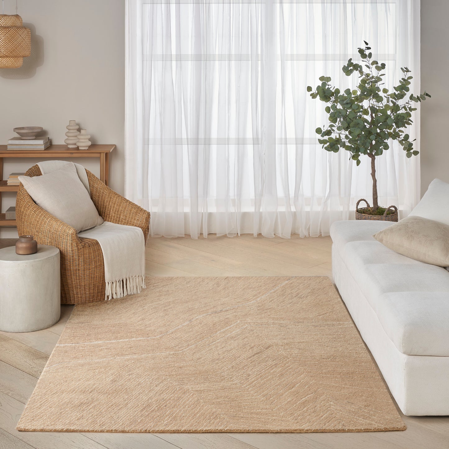 Bright neutral living room with beige area rug, white sofa, wicker armchair, potted tree, sheer curtains.