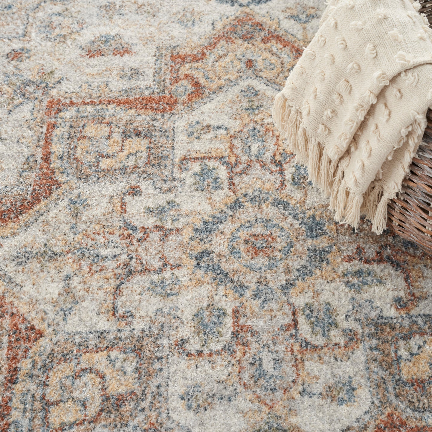 Vintage-style geometric patterned area rug in beige, blue and rust with a cream fringed throw over a wicker basket.