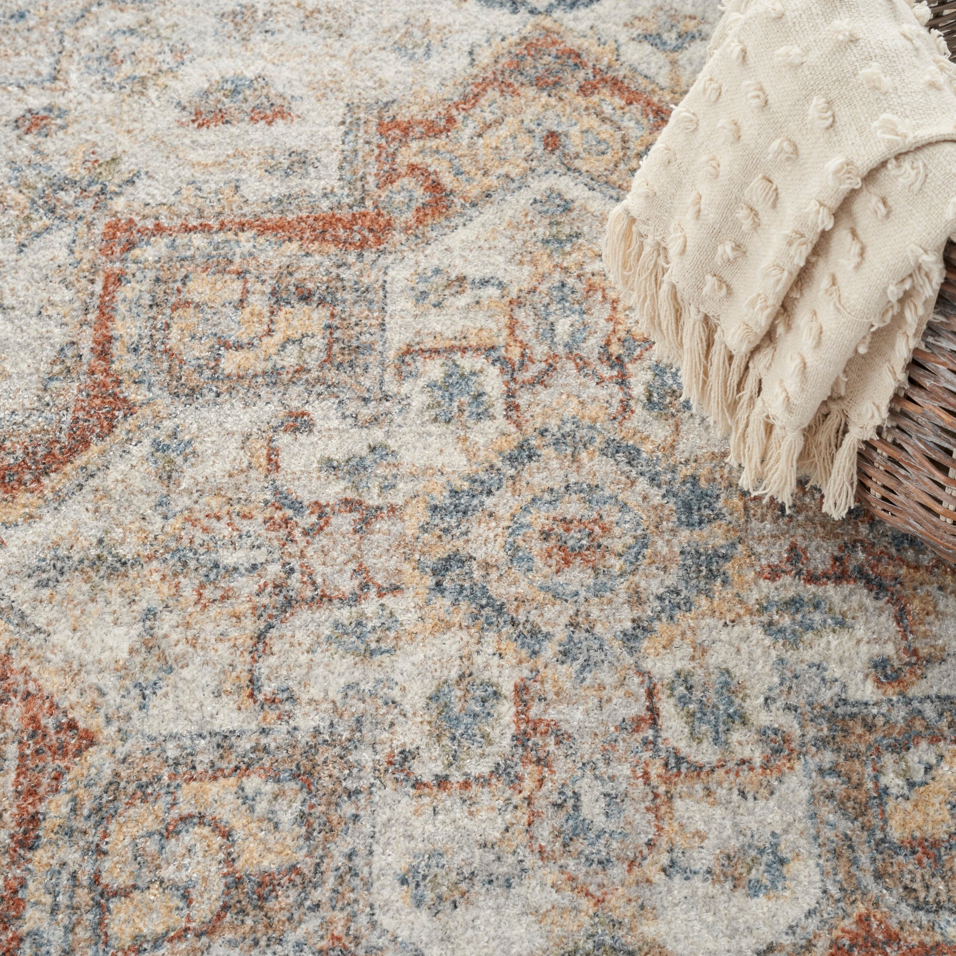 Vintage-style geometric patterned area rug in beige, blue and rust with a cream fringed throw over a wicker basket.