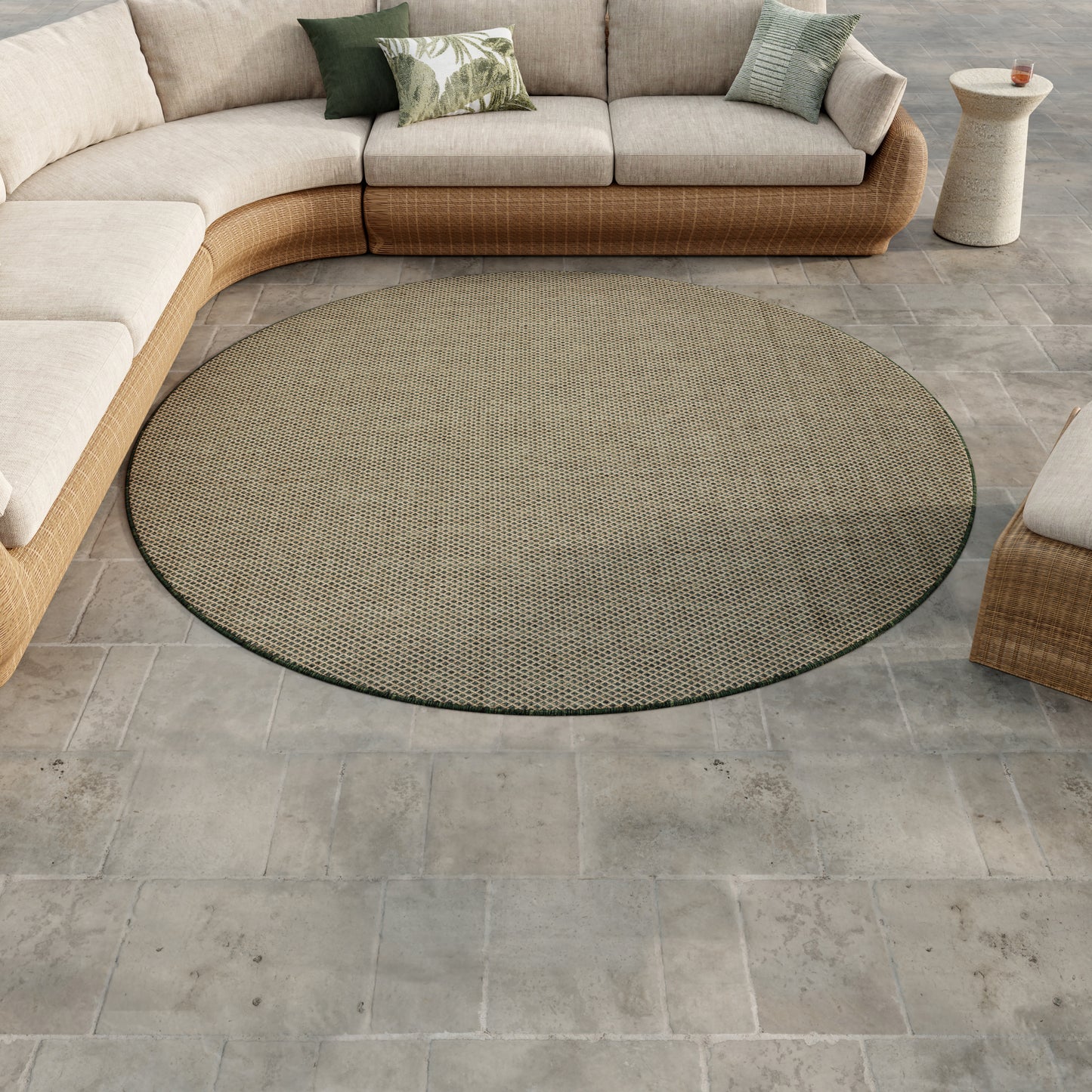 Round dark woven outdoor rug on gray tile patio, paired with beige wicker sectional and green cushions.