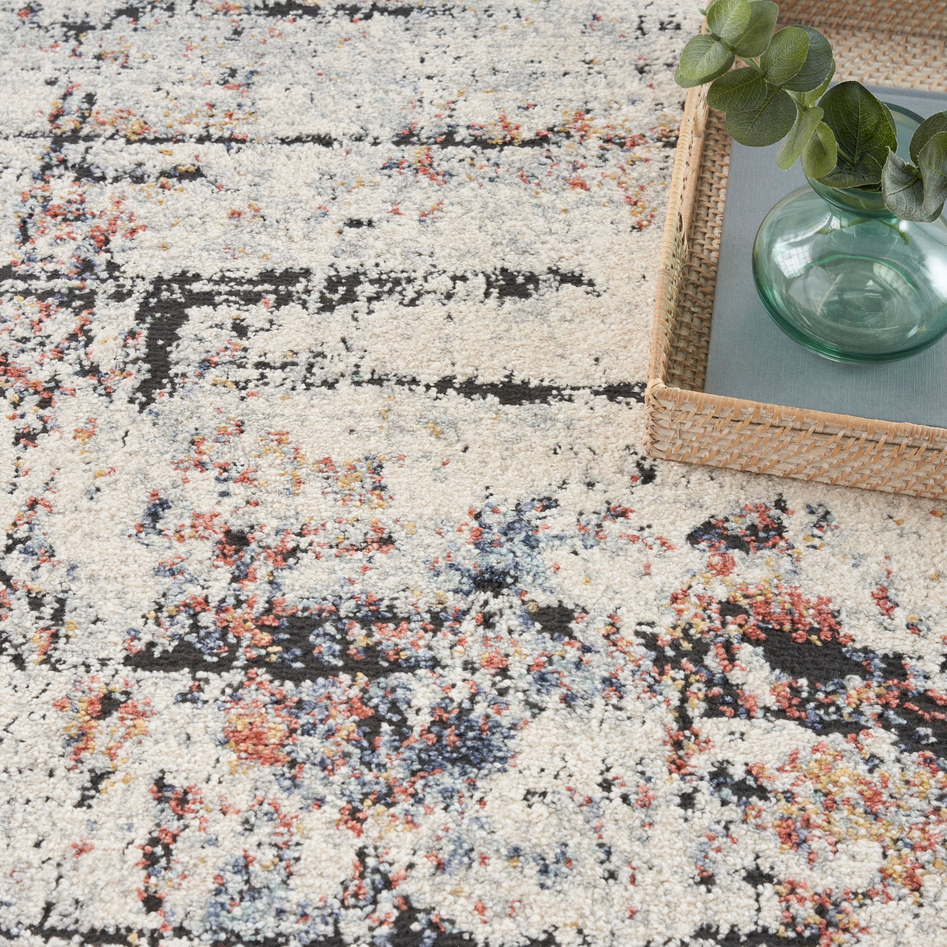 Beige abstract patterned rug with black and multicolor specks; wicker tray holding a green glass vase and eucalyptus leaves.