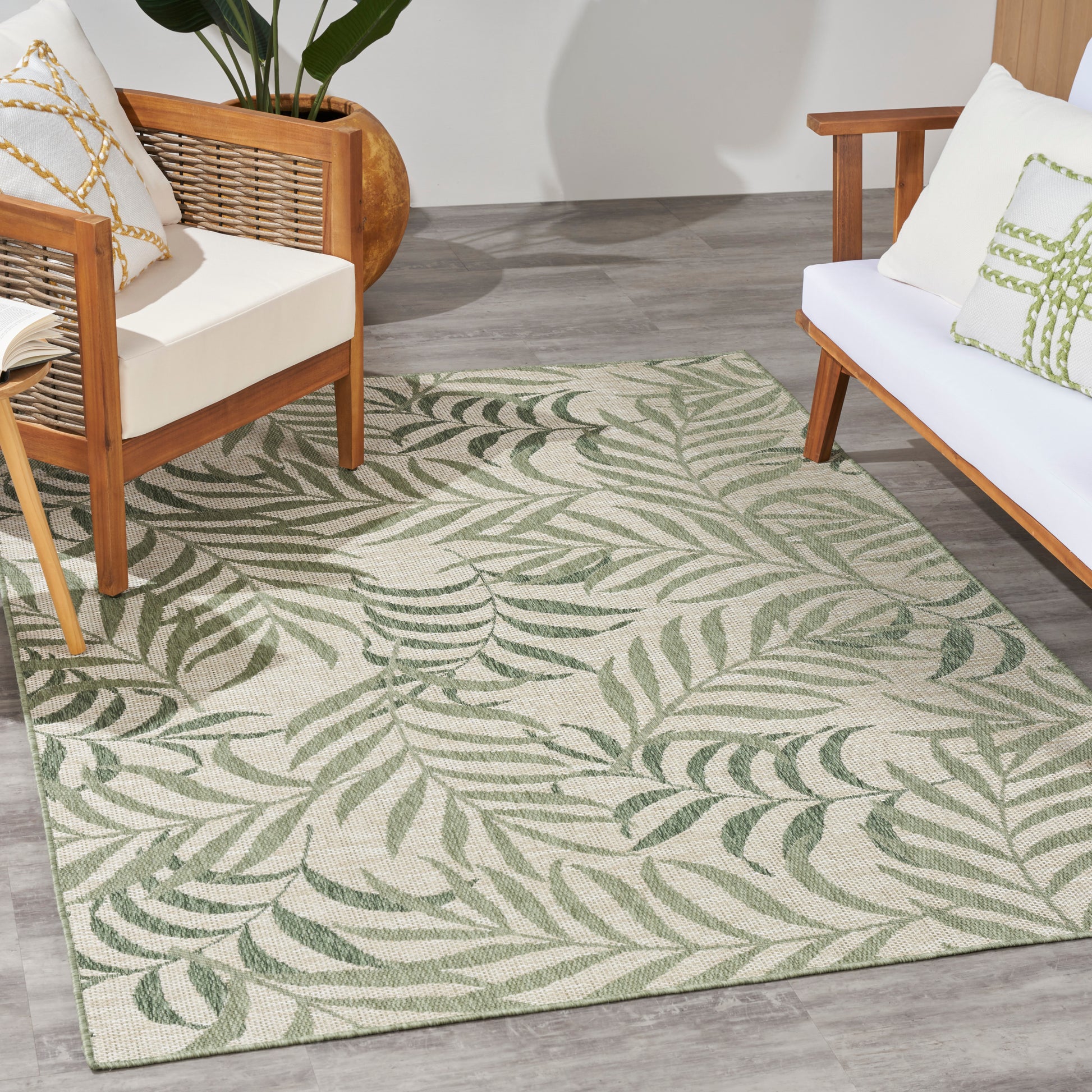 Green leaf-pattern outdoor area rug between mid-century wooden chairs with white cushions; potted plant nearby