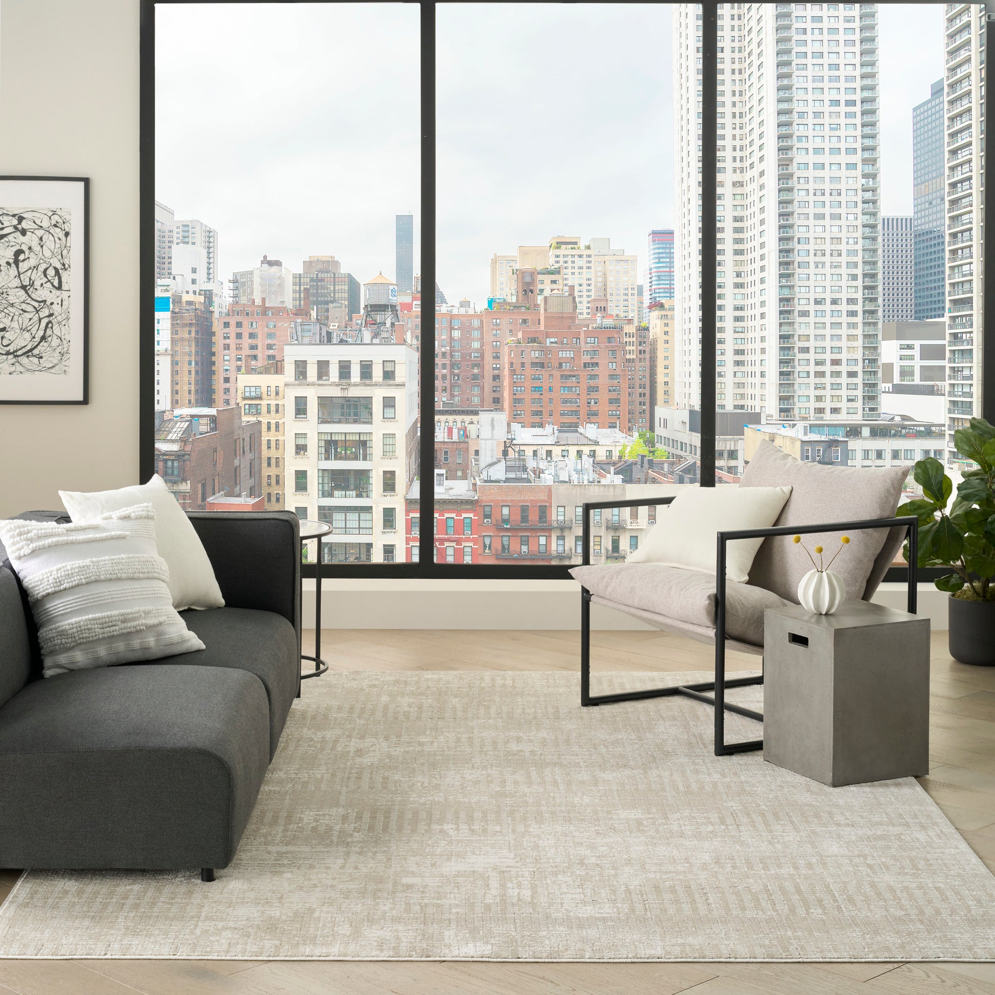 Modern living room with floor-to-ceiling city views, gray sofa, metal-framed armchair, concrete side table, and beige rug.