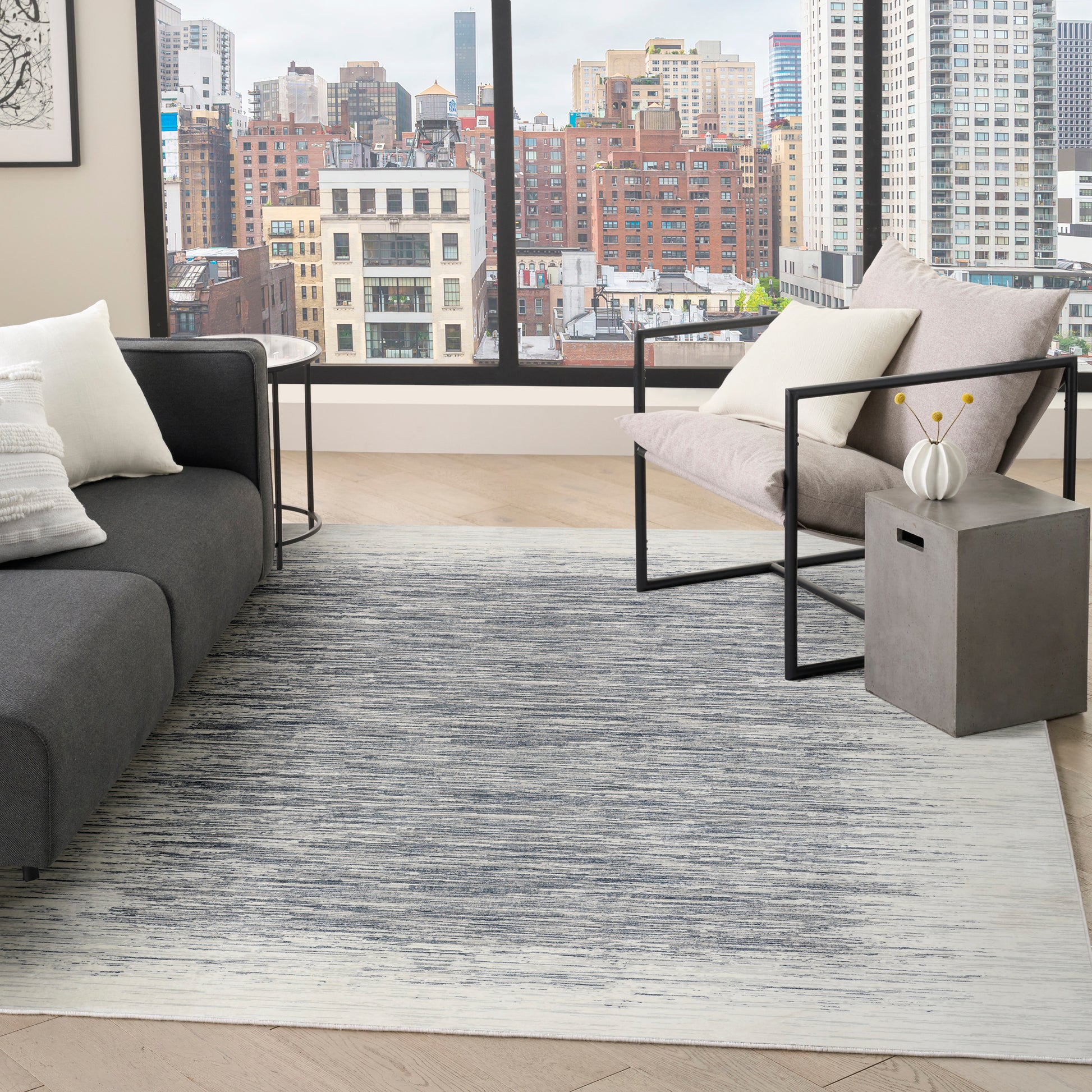 Modern living room with cityscape window, gray sofa, metal-framed chair, geometric rug, and concrete side table with white vase.