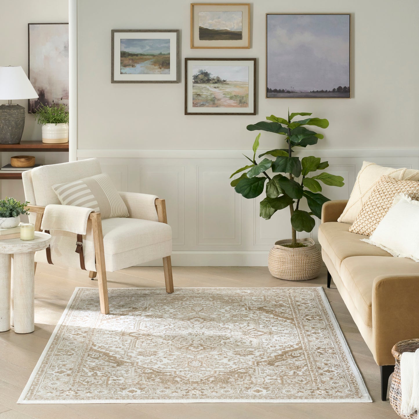 Neutral living room with beige sofa, cream armchair, woven patterned rug, potted plant, and landscape art gallery.