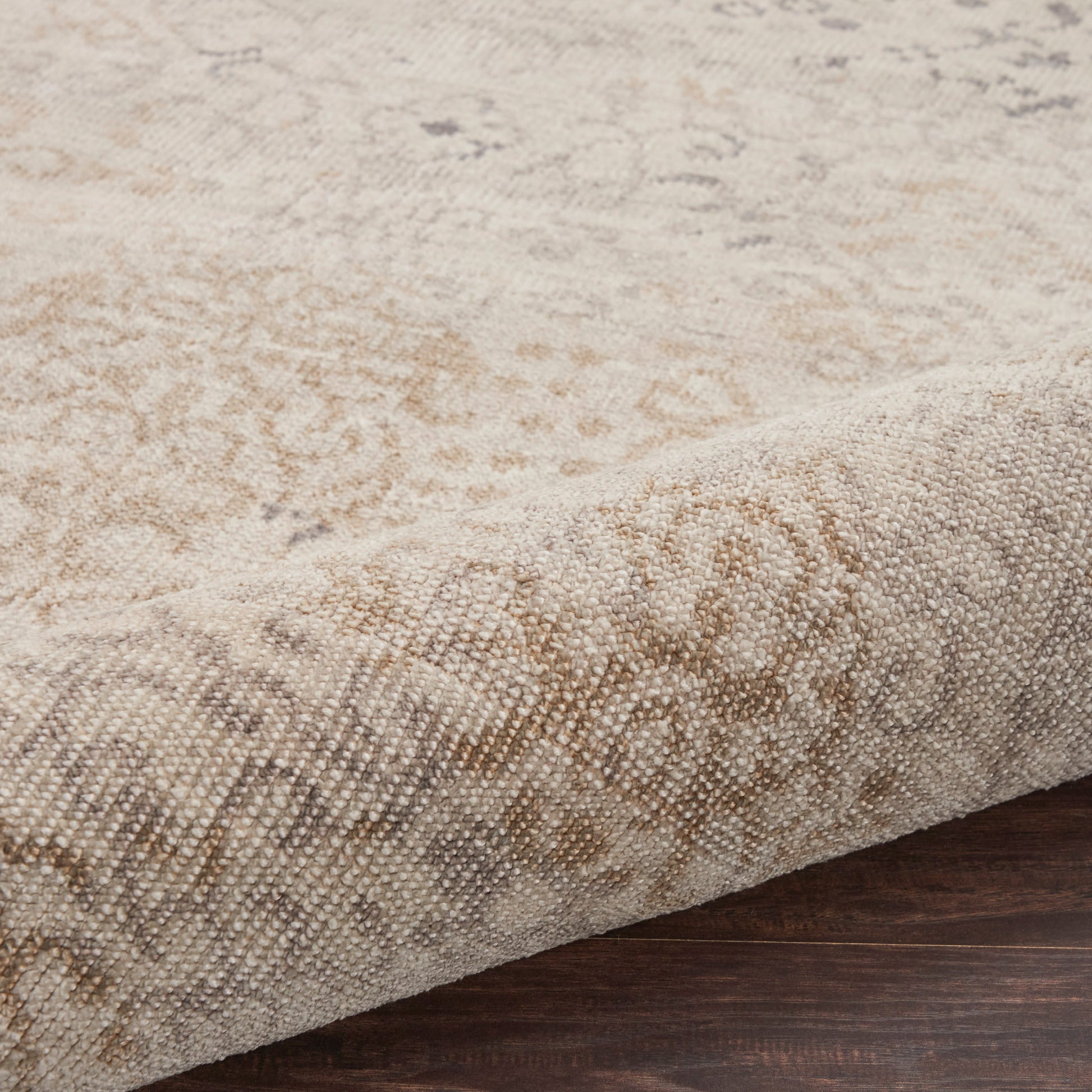 Beige patterned area rug with damask motif, textured woven pile & folded edge on dark wood floor.