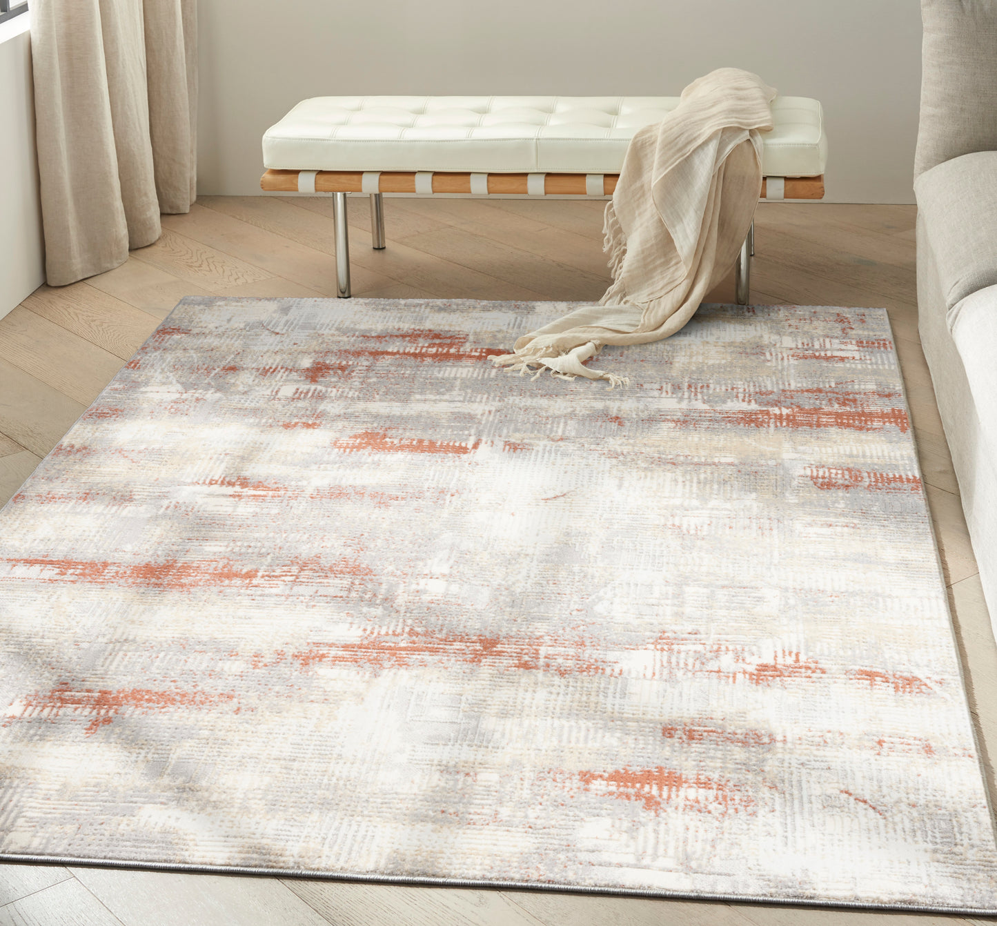 Abstract cream, gray and rust area rug on light hardwood floor beside a white tufted bench with wooden legs.