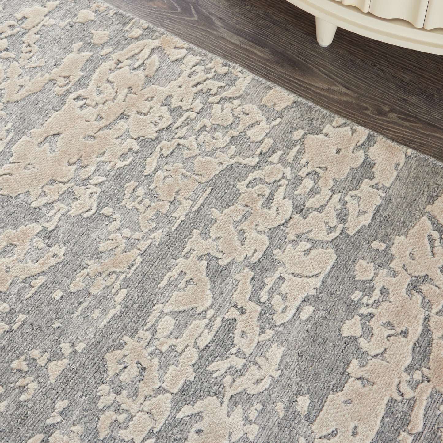 Gray and beige abstract-patterned area rug with a distressed look on a dark wood floor beside a cream-colored furniture leg.