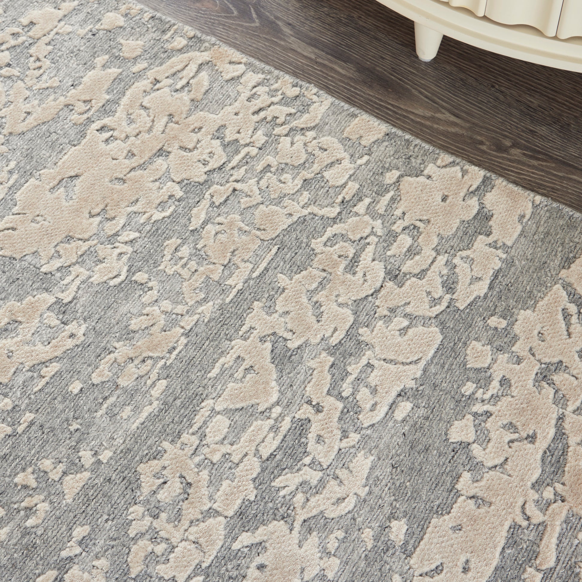 Gray and beige abstract-patterned area rug with a distressed look on a dark wood floor beside a cream-colored furniture leg.