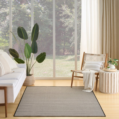 Bright living room with light wood floors, large windows, gray bordered rug, white sofa, wicker chair, plant, round ribbed side table.