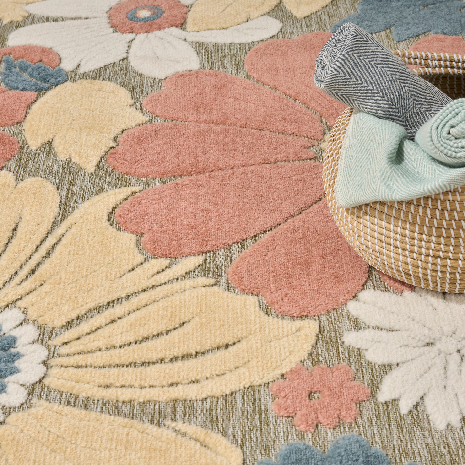Colorful floral area rug with large pink petals and yellow blooms; woven basket with rolled mint blanket and chevron towel.