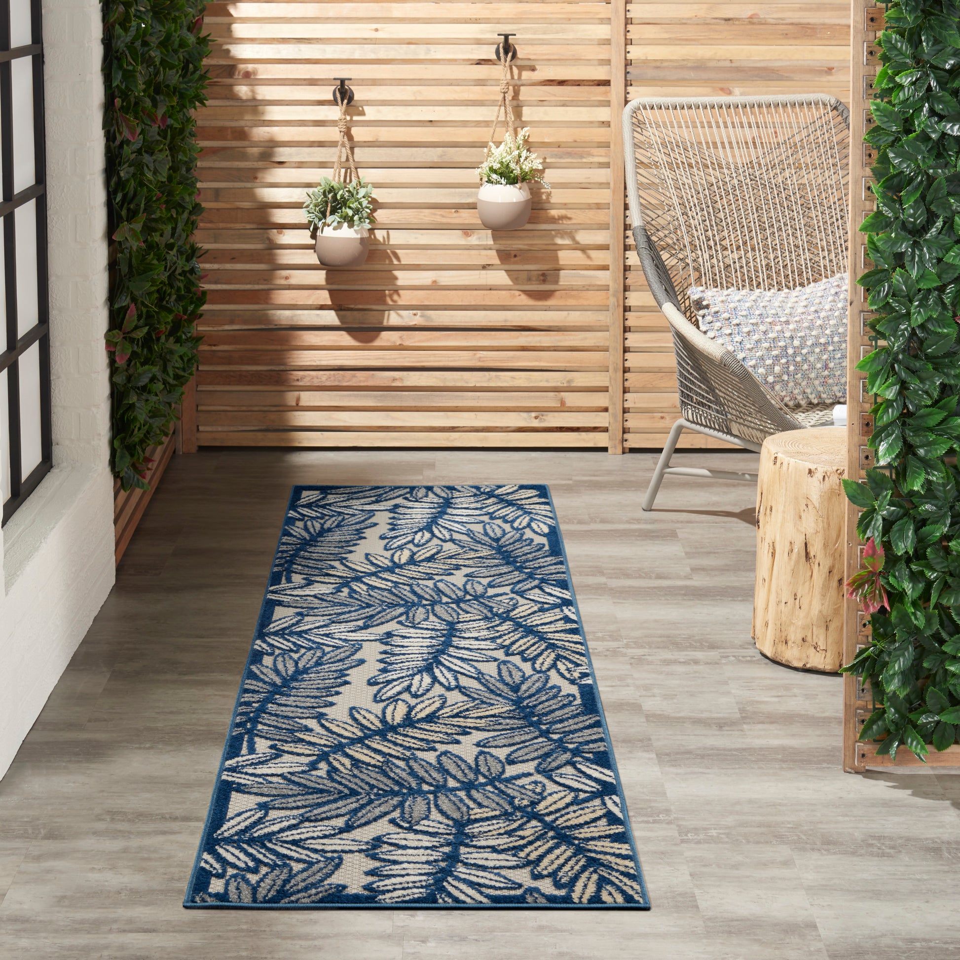Outdoor patio with blue fern-pattern rug, wooden slat privacy wall, hanging pots, wicker chair, and greenery.