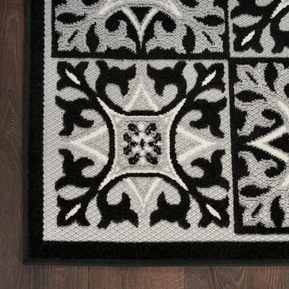 Black-and-white patterned rug with ornate scrollwork, square panel motifs, bordered in black on dark wooden floor.