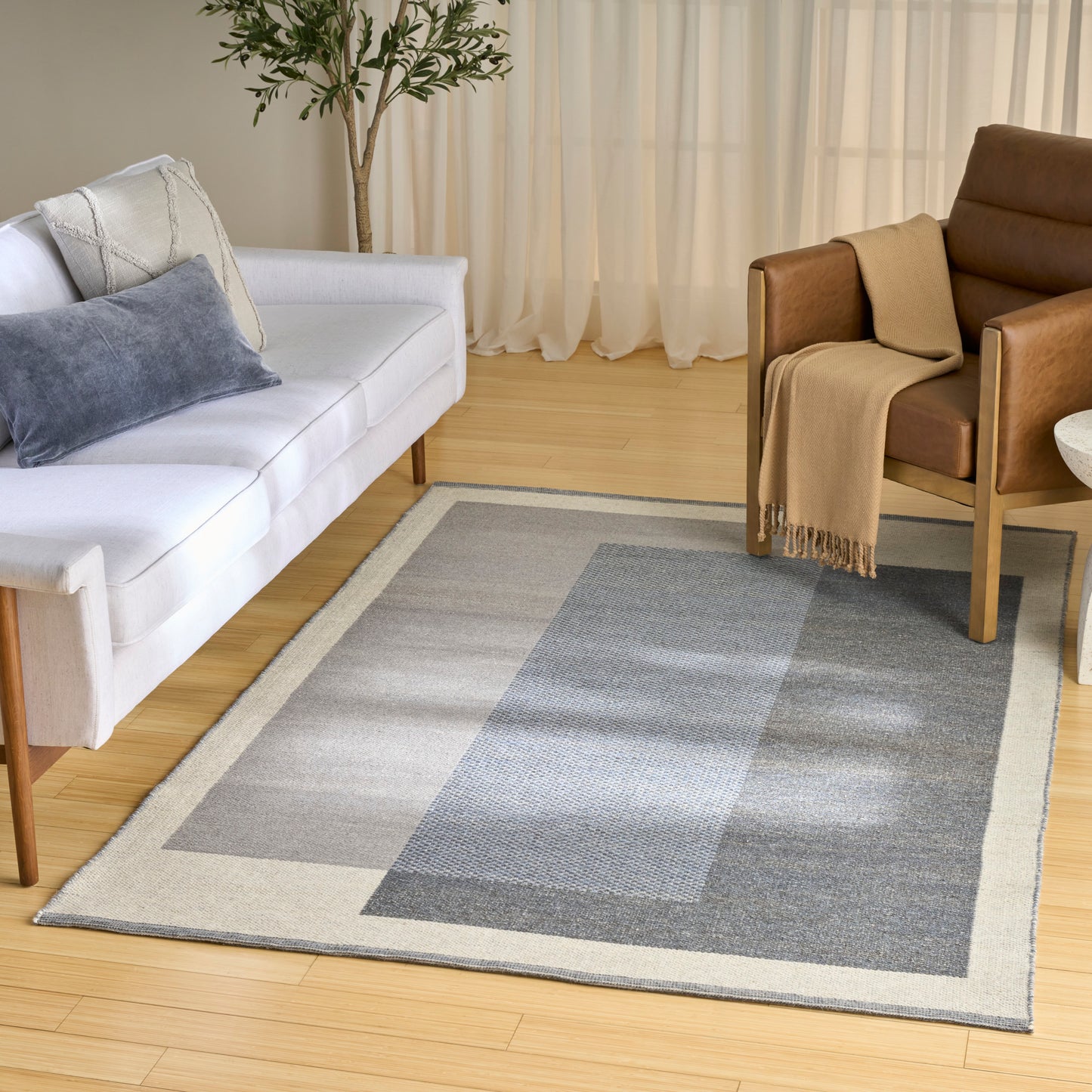 Rectangular geometric area rug in light gray and blue on wooden floor, white sofa, brown armchair, sheer curtains and plant.