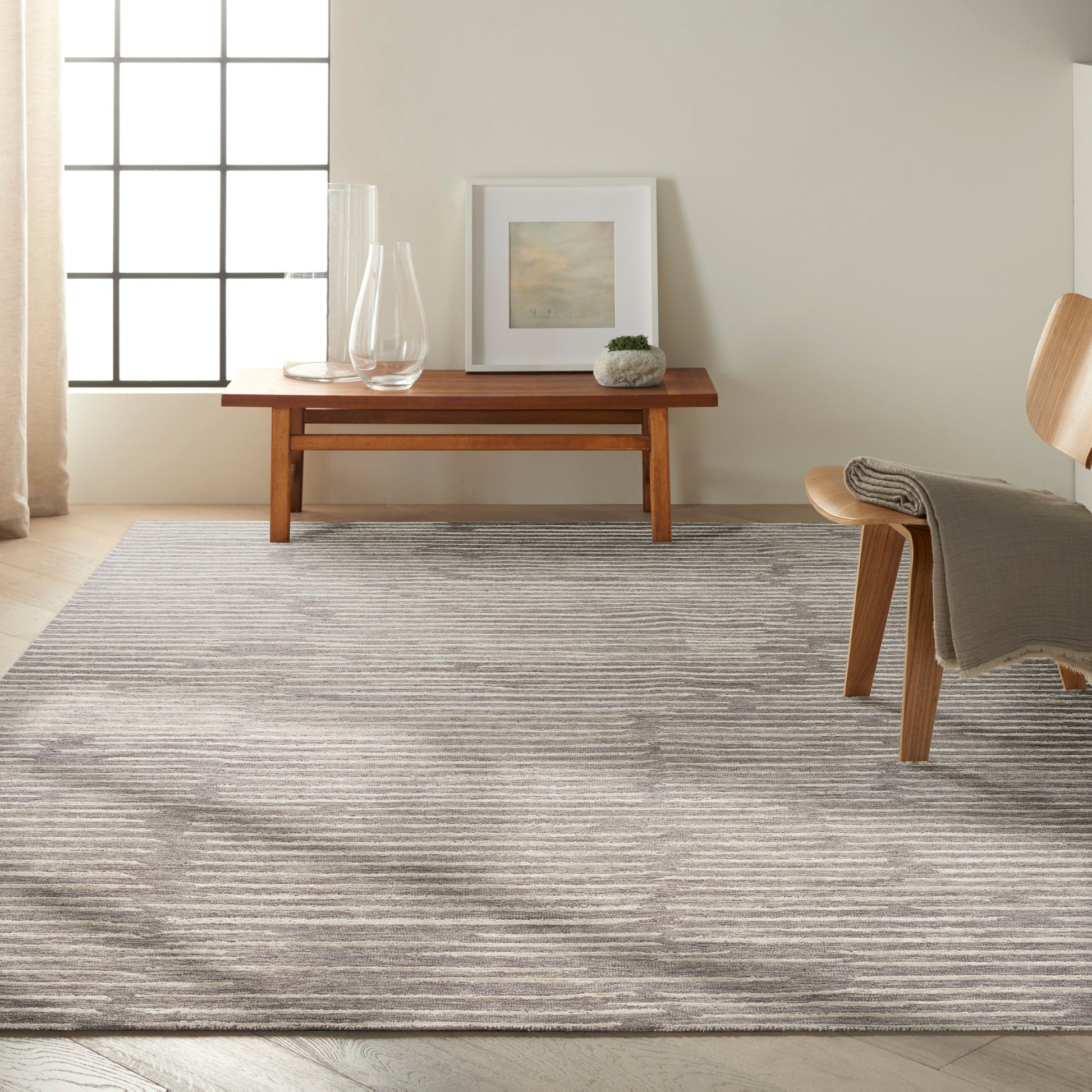 Light living room with a gray striped area rug, wooden coffee table, glass vases, framed art, and a wooden chair with a throw.