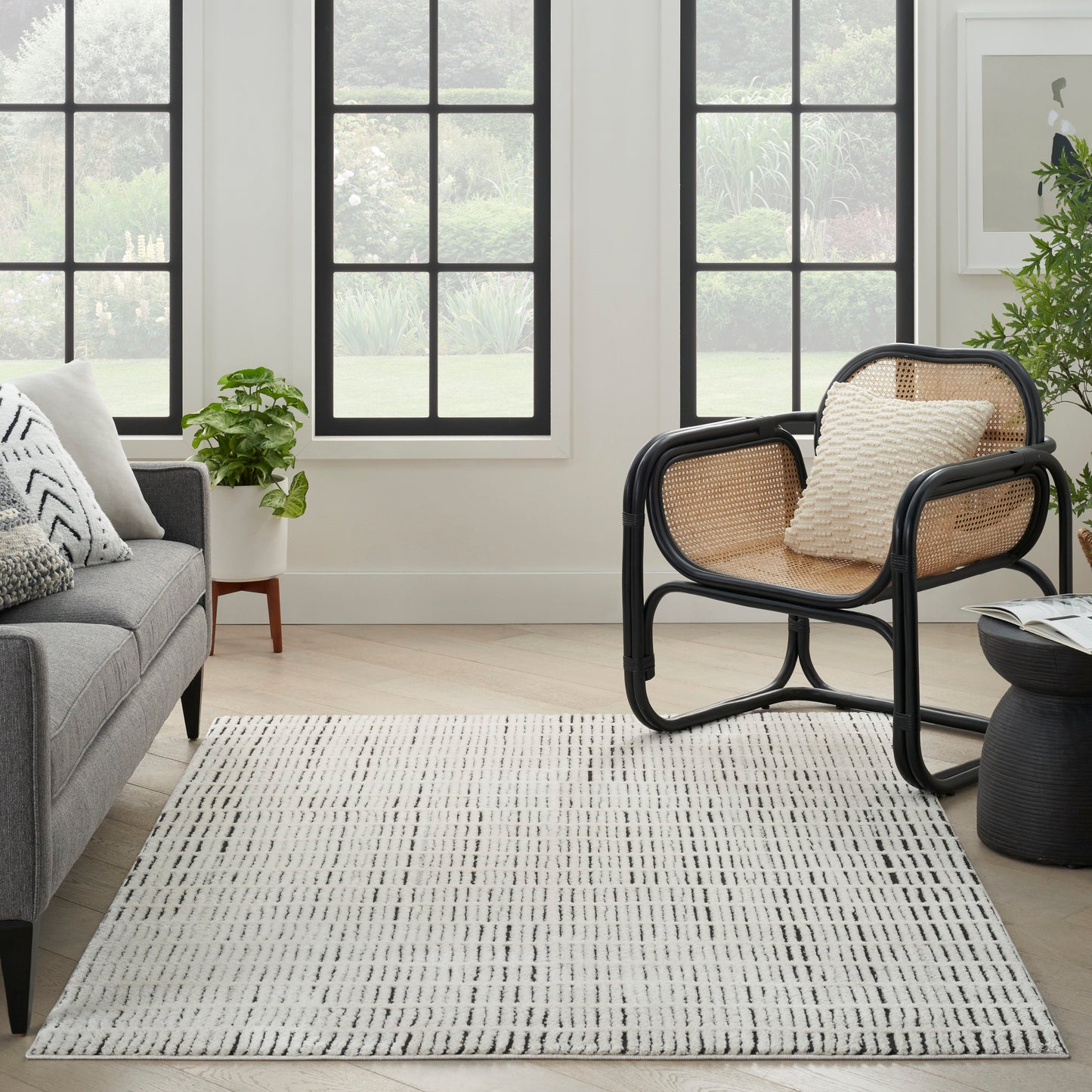 Living room with a black-rimmed cane armchair, beige cushions, gray sofa, striped light rug, potted plant, large windows.