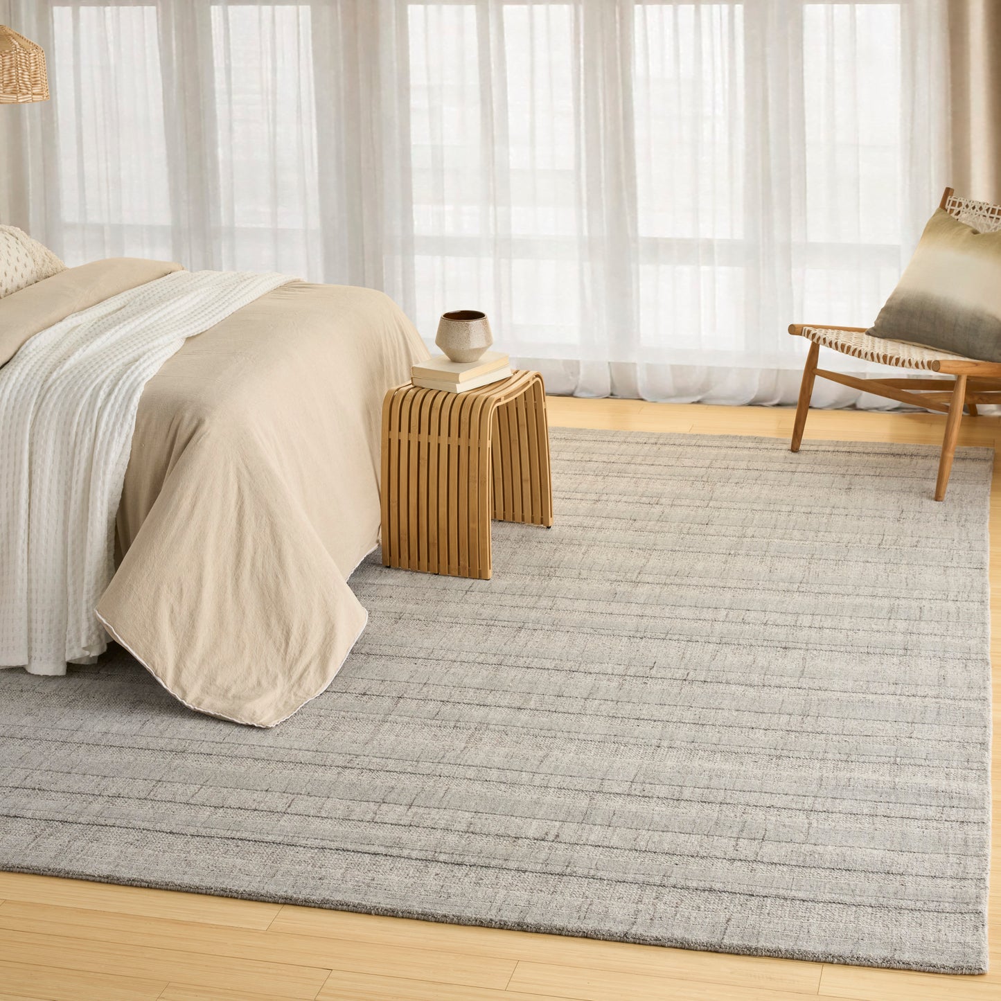 Neutral bedroom with beige duvet, light striped sheets, wooden side table, ceramic vase, woven chair, sheer curtains, rug.