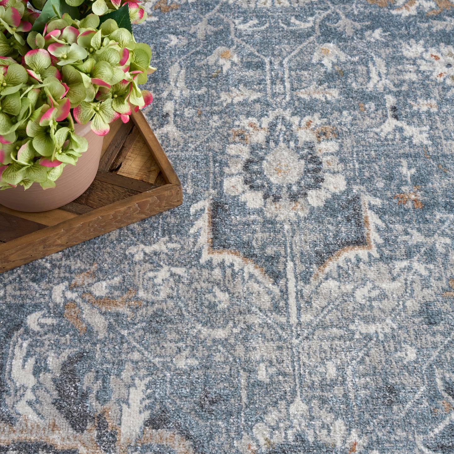 Blue-gray Oriental rug with faded ivory motifs; wooden tray holds a pink pot of green hydrangeas.