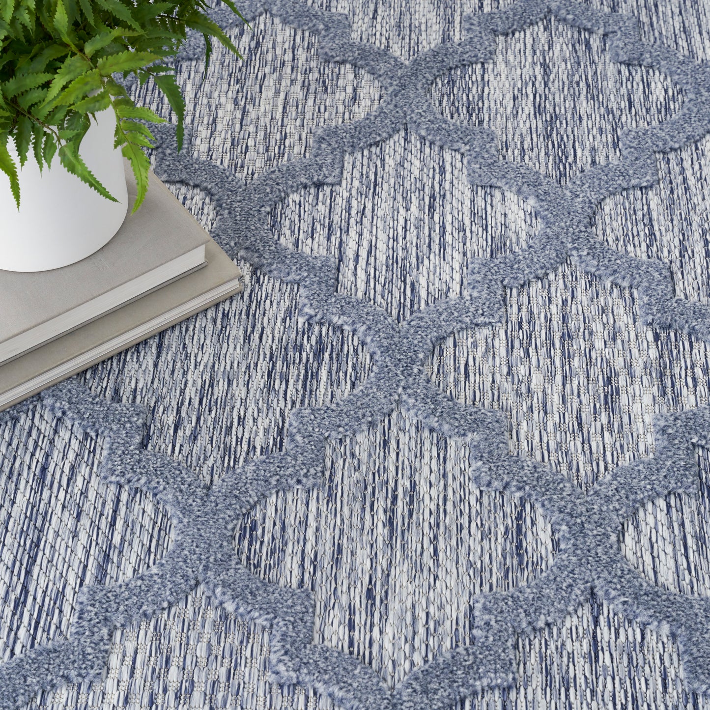 Blue-gray lattice-pattern rug with textured weave; white planter with green plant and stacked books nearby.