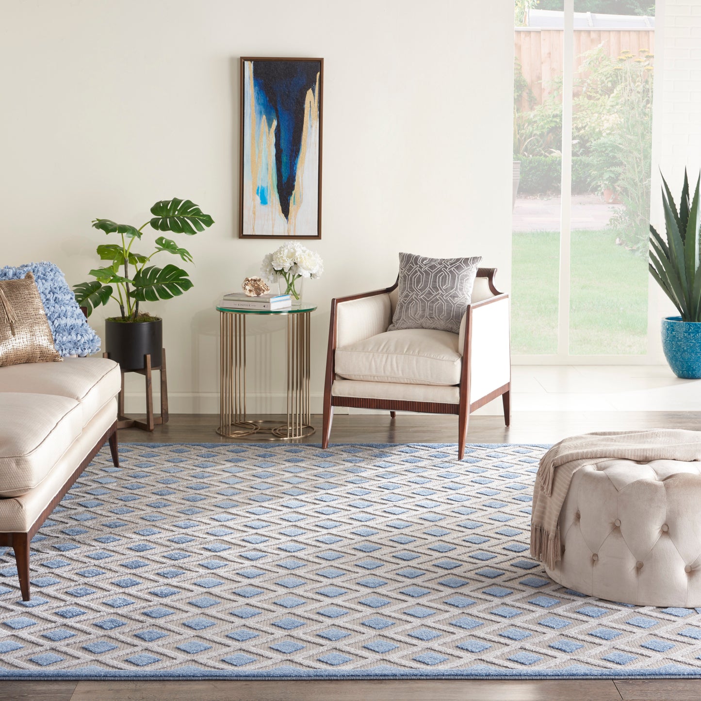 Cream living room with tufted ottoman, beige sofa, mid-century armchair, blue geometric rug, glass side table, abstract art.