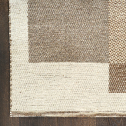 Rectangular woven area rug with cream border and taupe/brown patchwork blocks on dark wood floor.