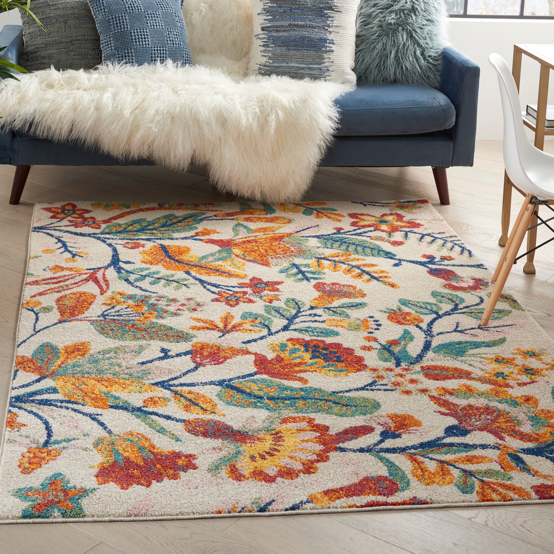 Colorful floral area rug with orange, blue, teal and red blooms on cream, in a living room with a navy sofa and fluffy throw.