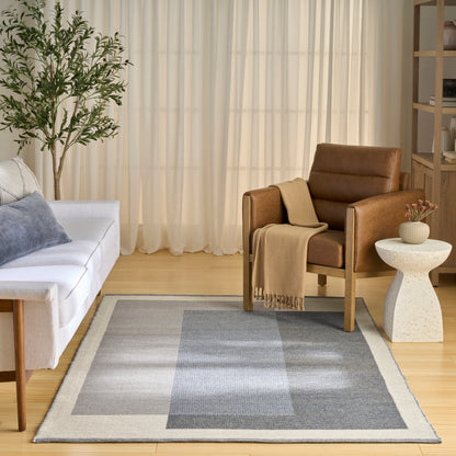 Cozy living room: brown leather armchair, white sofa, gray-blue rug, sheer curtains, plant, wooden bookshelf.