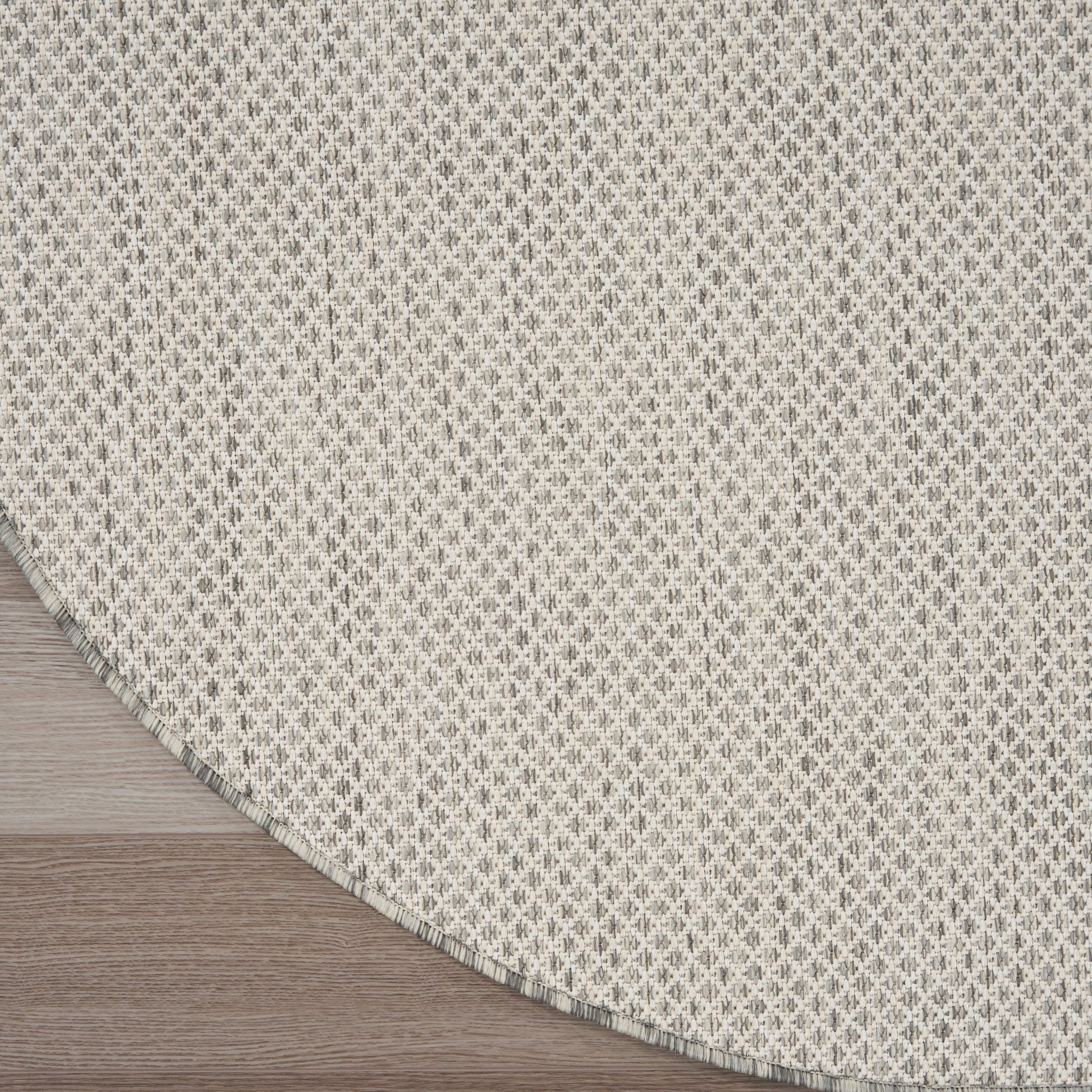Round beige woven rug with subtle grey lattice pattern and fringed edge on light wood floor.
