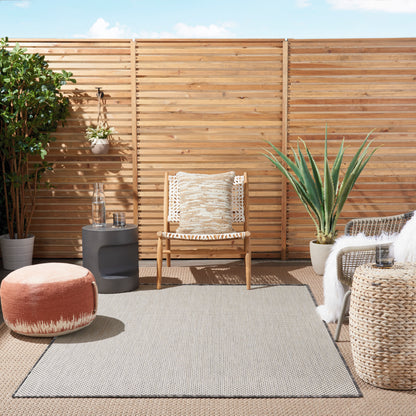 Outdoor patio with a gray modern rug as the focal item, alongside a woven armchair with beige cushion, an orange pouf, wicker tables, a potted agave, and a wooden slat fence.