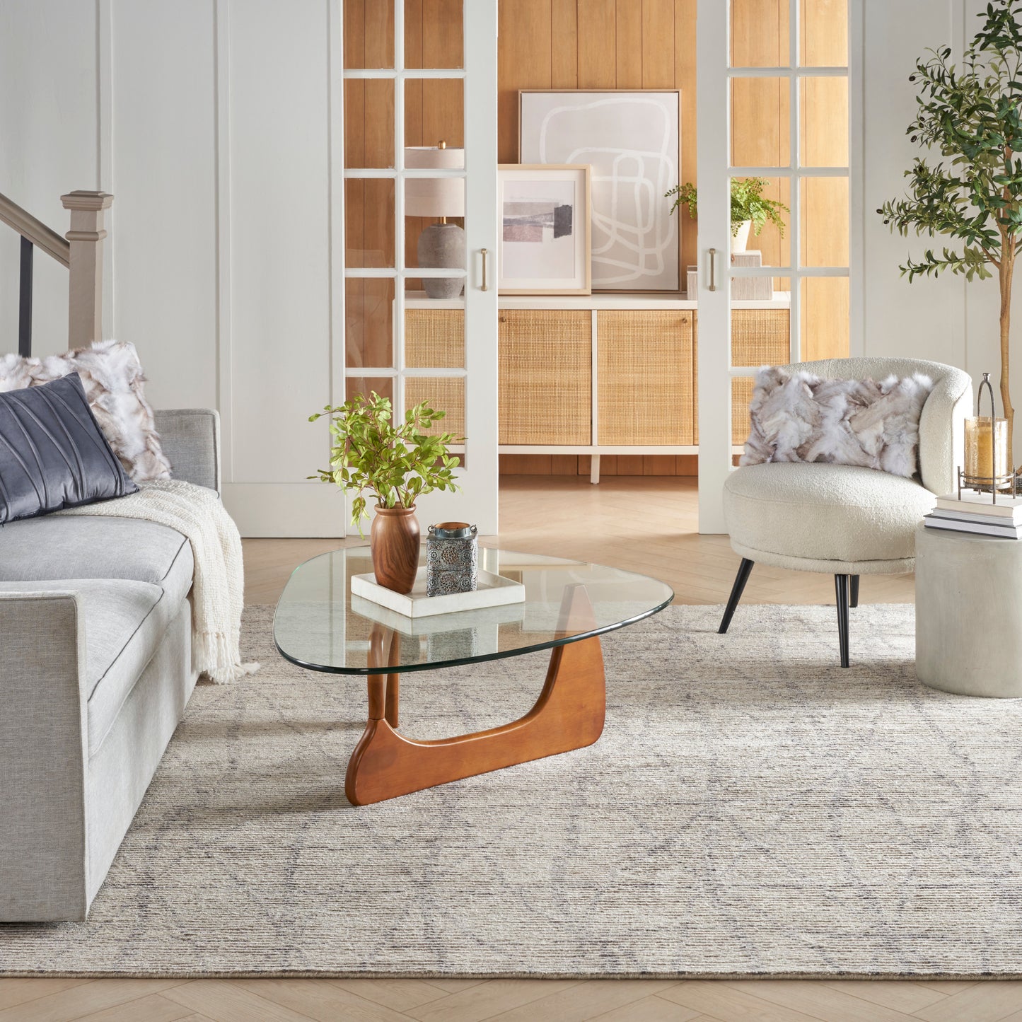 Round glass coffee table with sculptural orange wood base in a neutral living room with grey sofa and beige rug.