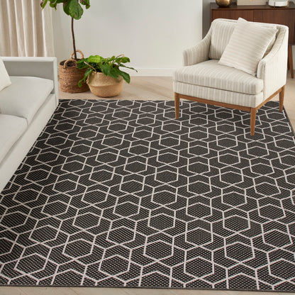 Dark gray geometric rug with white hex lattice, cream armchair on wooden legs, neutral living room with potted plants.