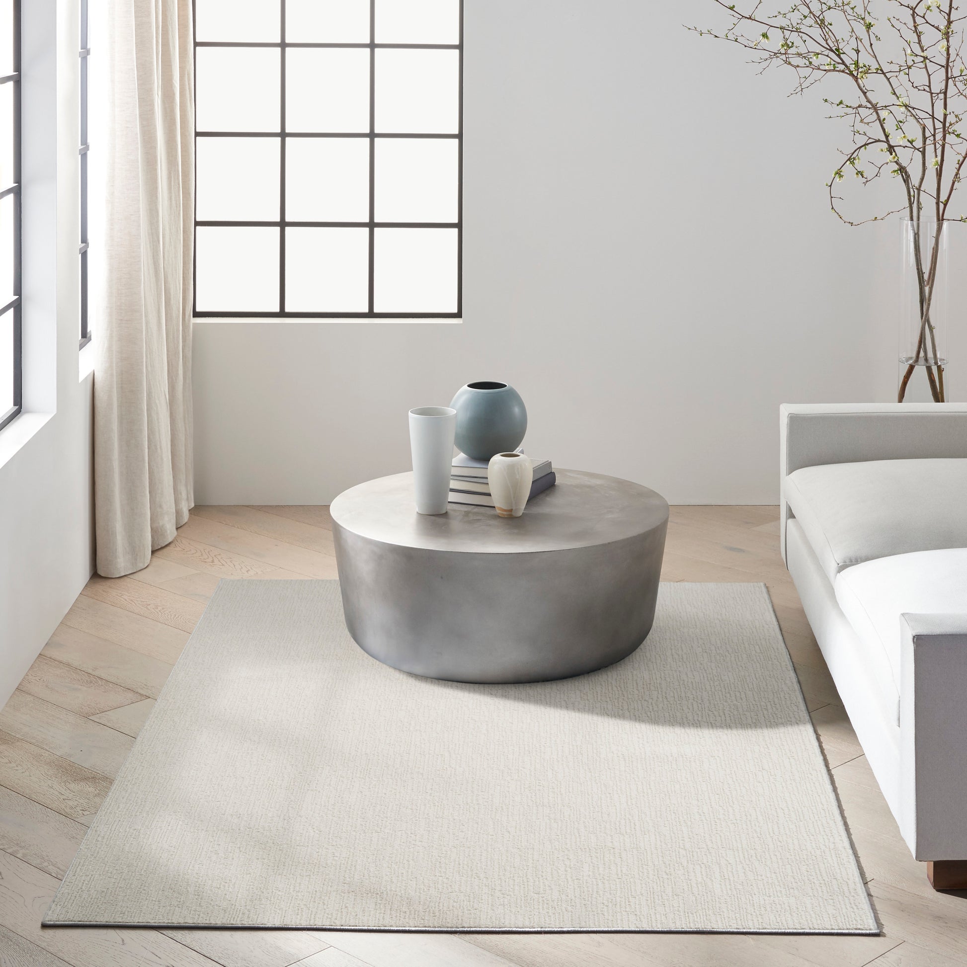 Minimalist living room with round concrete coffee table, neutral rug, white sofa, and decorative vases by a grid-window.