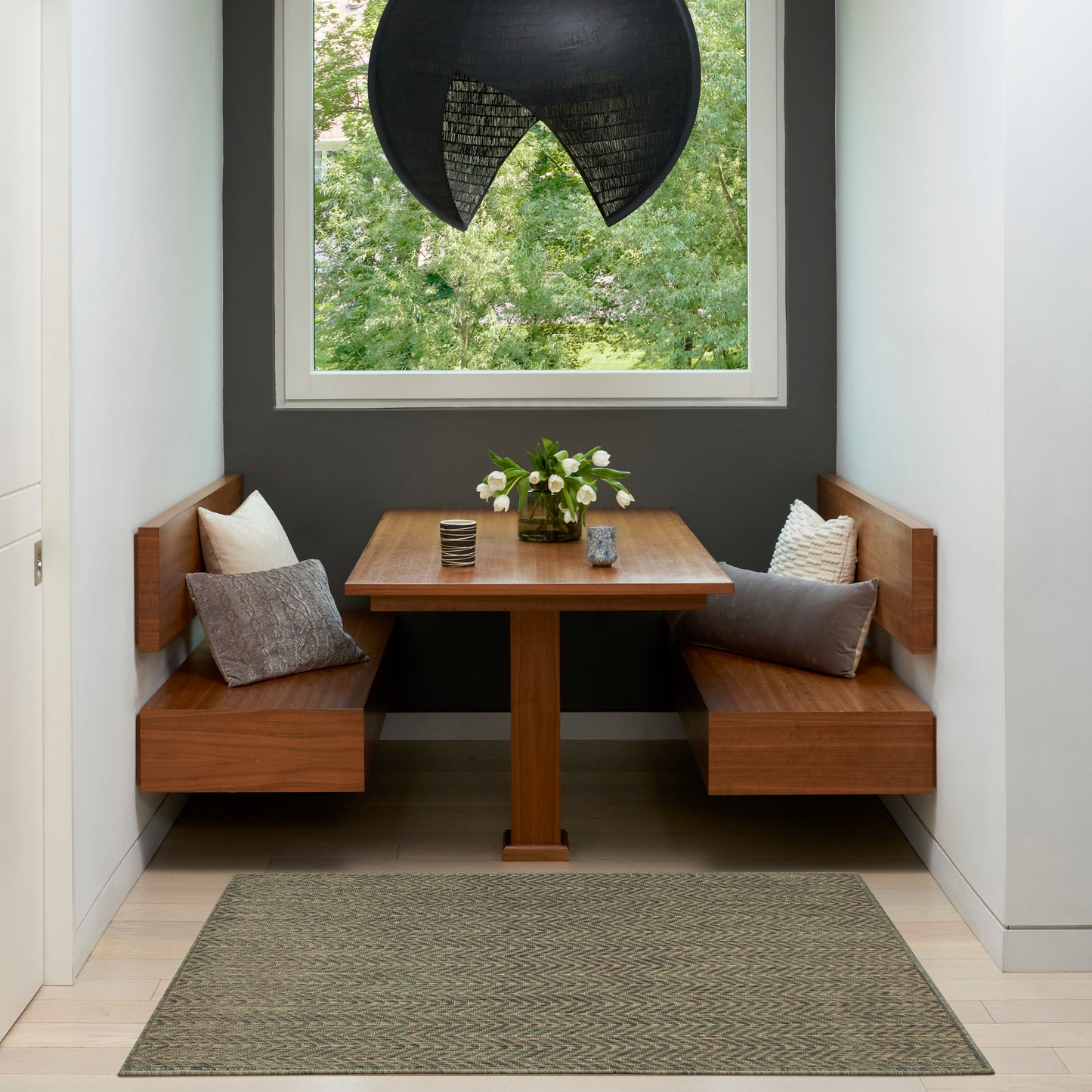 Built-in wooden booth dining nook with square table, gray walls, greenery outside window, black pendant light, cushions and vase of white flowers.