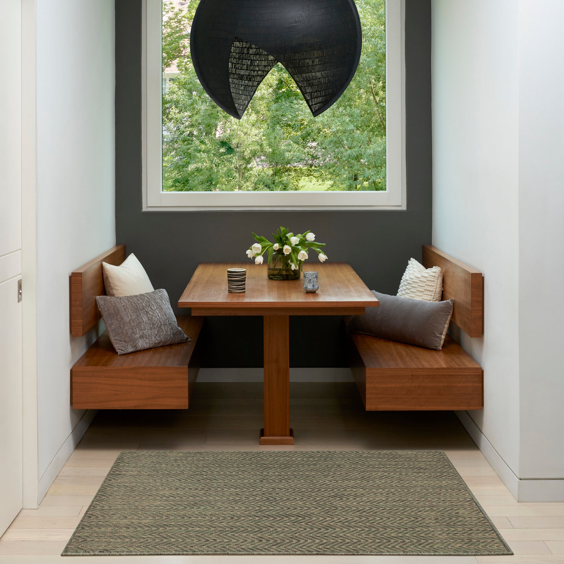 Built-in wooden booth dining nook with square table, gray walls, greenery outside window, black pendant light, cushions and vase of white flowers.