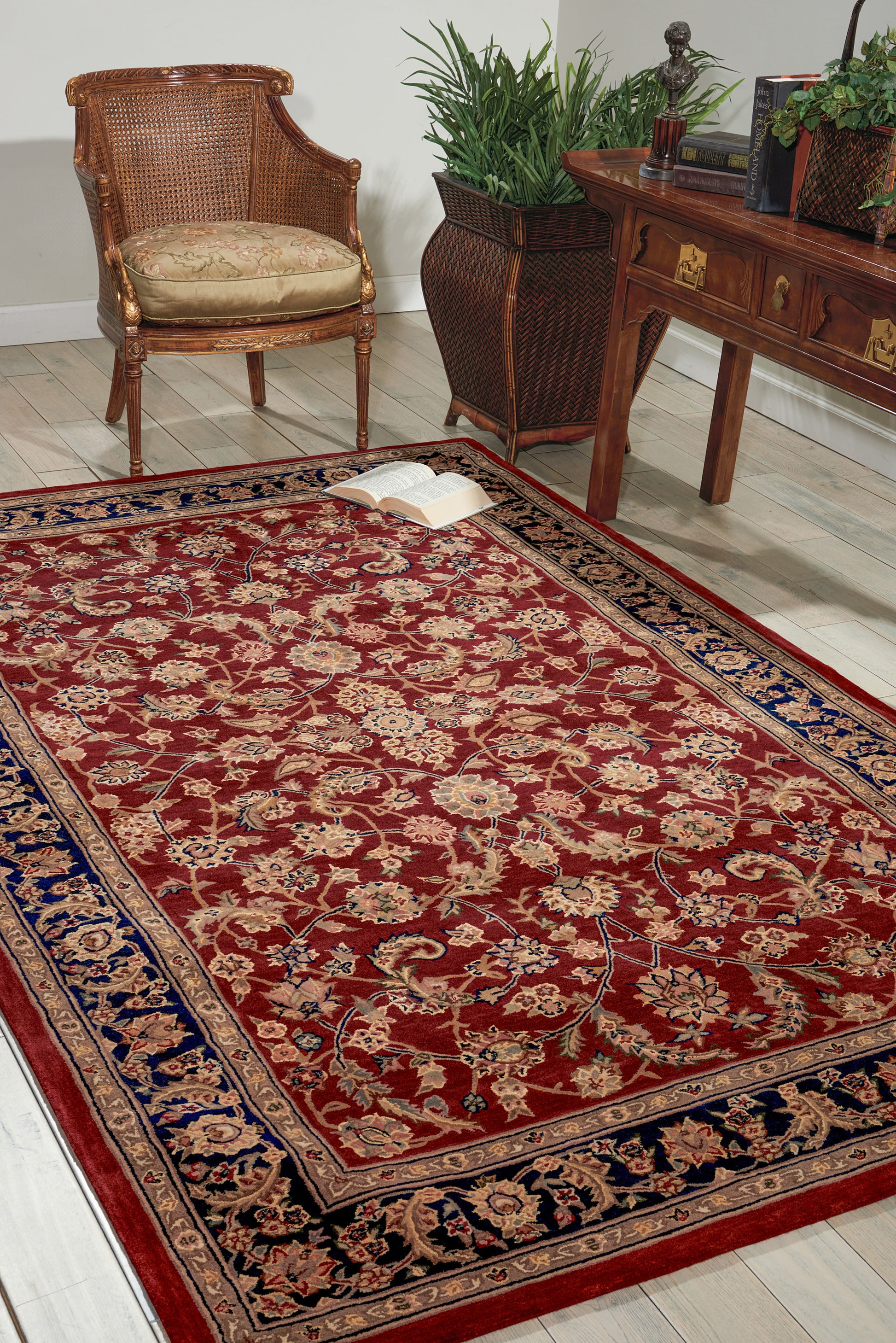 Red oriental rug with blue border in a living room with wicker chair, wooden desk, greenery, and an open book.